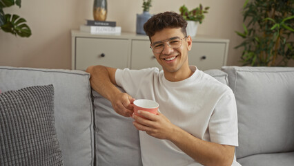 Fototapeta premium Handsome hispanic man smiling and holding a cup, wearing glasses and sitting on a couch in a cozy living room with indoor plants and light-colored decor.