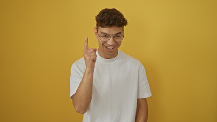 Young hispanic man in a white t-shirt with glasses pointing up and smiling over isolated yellow background wall