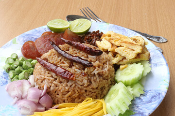 Rice mixed with shrimp paste consisting of sweet pork, chinese sausage, fried eggs, cucumber slice, sour mango, shallot, yard long bean, roasted peppers and lemon. Placed with a spoon and fork.
