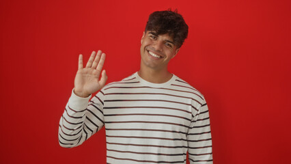 Naklejka premium Handsome young hispanic man smiling and waving in a striped shirt against an isolated red background.