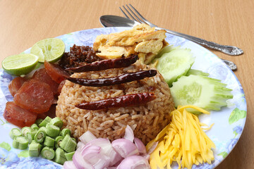 Rice mixed with shrimp paste consisting of sweet pork, chinese sausage, fried eggs, cucumber slice, sour mango, shallot, yard long bean, roasted peppers and lemon. Placed with a spoon and fork.
