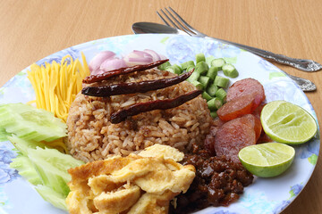 Rice mixed with shrimp paste consisting of sweet pork, chinese sausage, fried eggs, cucumber slice, sour mango, shallot, yard long bean, roasted peppers and lemon. Placed with a spoon and fork.
