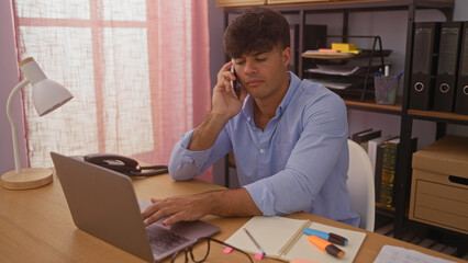 Handsome hispanic man working in an office while talking on the phone using a laptop and taking notes.