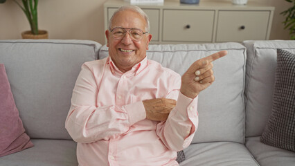 Middle-aged caucasian man smiling and pointing while sitting in a cozy living room, enjoying a relaxed moment indoors.