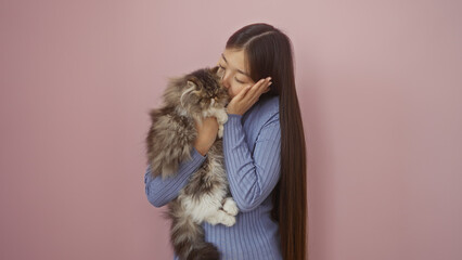 A young chinese woman cuddles her furry cat against a pink background, showcasing a loving and tender bond between the pet and owner.