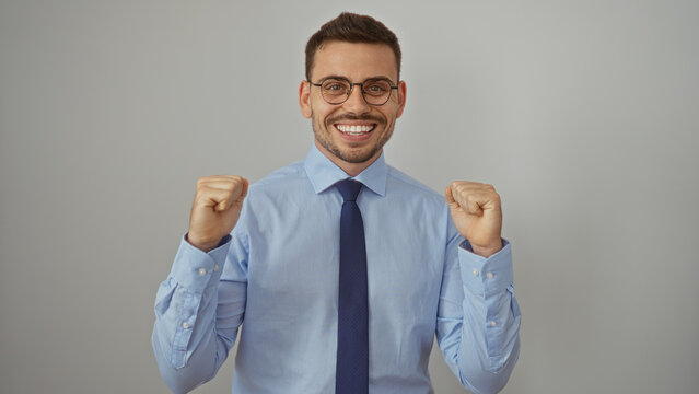Young hispanic man with a beard in a blue shirt and tie, smiling confidently with fists raised, standing isolated against a white background.