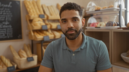 Young hispanic man in a bakery shop with fresh bread and pastries, smiling at the camera, showcasing a cozy and welcoming indoor environment for customers.