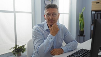 Middle aged man with glasses seated in office with hand on chin, wearing blue shirt, with plant and computer on desk next to window