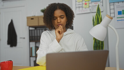 Young african american woman with curly hair, thinking deeply while working on a laptop in a modern indoor office environment.