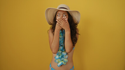 Young woman wearing a summer hat and blue lei covers her mouth with her hands in surprise against...