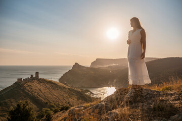 A woman stands on a rocky hill overlooking the ocean. She is wearing a white dress and she is enjoying the view. The scene is serene and peaceful, with the sun shining brightly in the background.