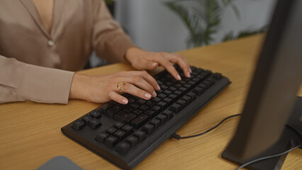 A woman types on a keyboard in an office setting, showcasing her hands with a gold ring, surrounded by indoor plants and a wooden desk.