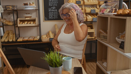 Mature grey haired woman smiling in a bakery shop surrounded by fresh bread and pastries, engaging with a laptop