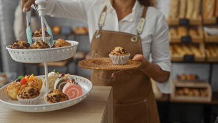 Woman working in a bakery holding a tray of muffins and pastries while wearing an apron indoors
