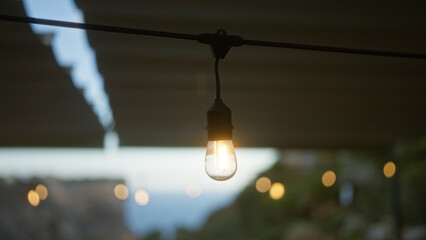 Hanging light bulb glowing outdoors with blurred bokeh lights in the background, creating a serene evening ambiance.