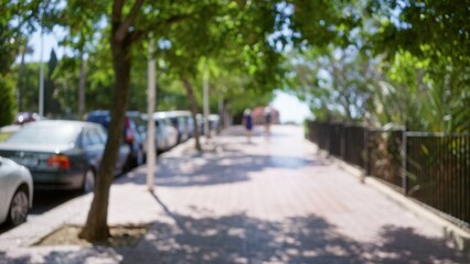 Naklejka premium Defocused view of an outdoor path lined with parked cars and shaded by trees with blurred people in the background