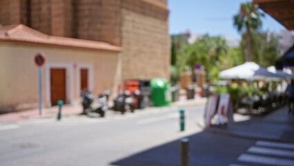 Fototapeta premium Blurred outdoor street scene with motorcycles, buildings, and defocused people under umbrellas in a bright, sunny day