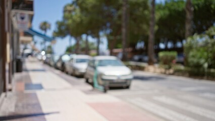 Defocused outdoor scene on a sunny day with blurred street view featuring parked cars, trees, and a sidewalk indicating urban life.