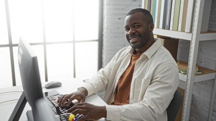Young african man working on computer in an office setting, smiling confidently with shelves and windows in the background