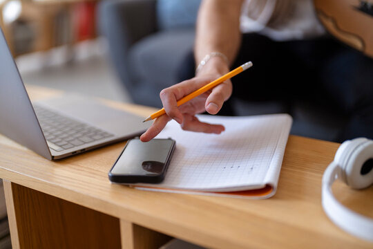 Close-up shot of a hand holding a pencil over a notebook, with an oud nearby, capturing the intimate moment of musical creation and notation in a quiet, comfortable indoor space.