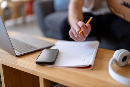 Close-up shot of a hand holding a pencil over a notebook, with an oud nearby, capturing the intimate moment of musical creation and notation in a quiet, comfortable indoor space.