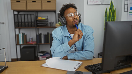 Young african american man with a beard and glasses, wearing headphones, sitting at a desk in an office, pensively looking at a computer screen with a pen in hand, surrounded by books and files.