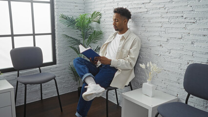 A young, handsome, african american man with a beard sits reading a book in an indoor waiting room with modern decor and a large window, creating a peaceful ambiance.