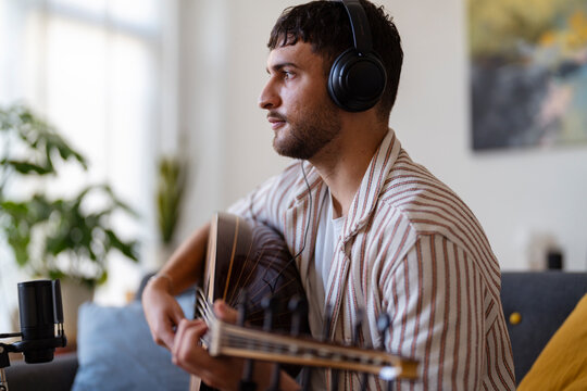 A young Arab man with headphones plays an oud in a relaxed, modern living room, focusing on recording his music, showing dedication to his craft and concentration in an inspiring environment.