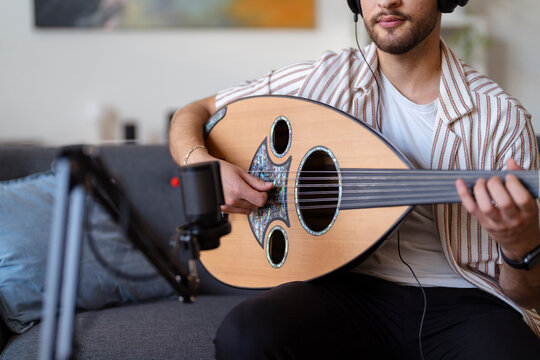 Close-up of a man wearing headphones, strumming an oud while recording in a cozy living room, capturing the essence of home music production and passion for musical creation.
