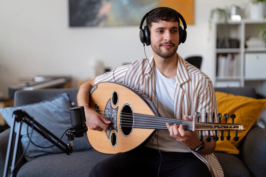 Close-up of a man wearing headphones, strumming an oud while recording in a cozy living room, capturing the essence of home music production and passion for musical creation.