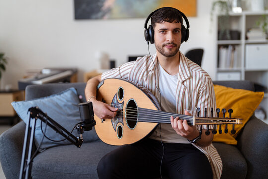 Close-up of a man wearing headphones, strumming an oud while recording in a cozy living room, capturing the essence of home music production and passion for musical creation.