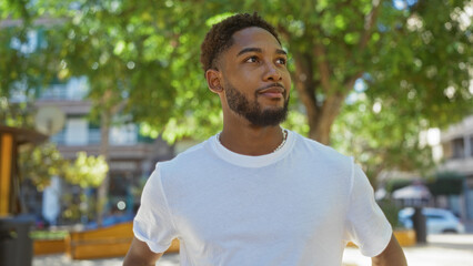 Handsome young man with a beard in a white shirt standing in an urban park with trees and buildings in the background on a sunny day