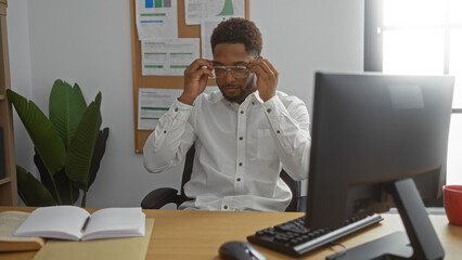 Young african american man adjusting glasses in office room with desk and computer