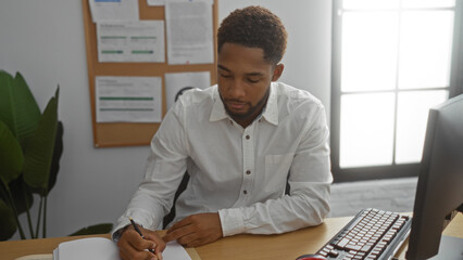 Young man working in an office, writing on a paper with a pen, seated in front of a computer, with...