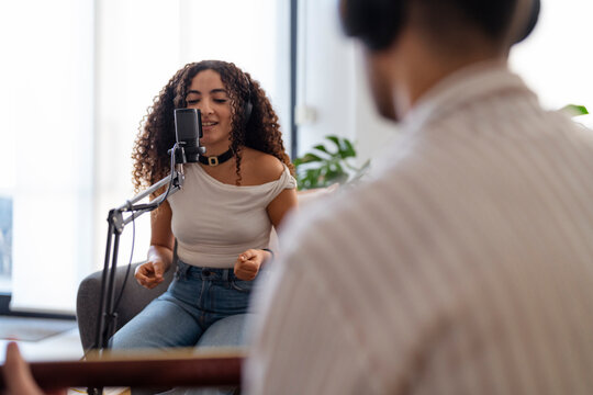Arab woman sings into a microphone while a man plays the guitar, both engaging in a creative music recording session in a cozy, well-decorated living room, capturing teamwork and musical passion.
