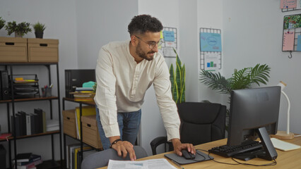 A handsome young hispanic man working in a modern office, leaning over a desk while using a computer, surrounded by indoor plants and organized shelves.