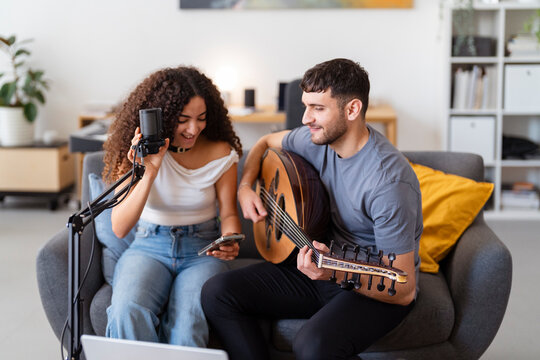 Arab man with an oud and a woman with a microphone share a music session on a cozy couch, capturing their enjoyment and creative energy in a relaxed home studio environment.
