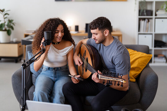 Arab man with an oud and a woman with a microphone share a music session on a cozy couch, capturing their enjoyment and creative energy in a relaxed home studio environment.