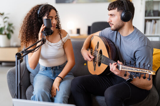 A young man plays an oud while a woman sings into a microphone, both wearing headphones, engaging in a focused recording session in a cozy home setting emphasizing creativity and teamwork.
