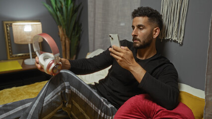 Young hispanic man takes a photo of headphones with his phone while sitting on a bed in a cozy bedroom at home.