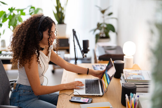 Arab young woman wearing headphones edits audio on a laptop in a cozy home studio, with books and a microphone nearby, embodying a creative and focused workspace for audio production.