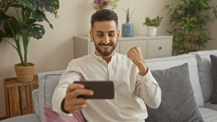 Young hispanic man in a living room smiling at his smartphone while sitting on a couch with indoor plants in the background.