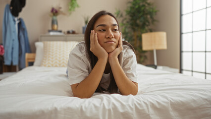 Young, attractive, hispanic woman lying on a bed in a cozy bedroom interior, appearing thoughtful and relaxed with a homey atmosphere.