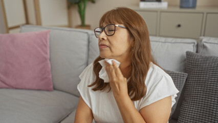 Mature hispanic woman experiencing a hot flash, sitting in her living room, wiping sweat with a tissue.