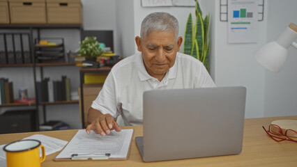 Elderly hispanic man working in an office on a laptop with documents and a coffee cup on the desk, demonstrating a professional indoor work environment.