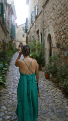 Woman walking on a cobblestone street in valldemossa, mallorca, spain, wearing a green dress, surrounded by rustic stone buildings with green shutters.