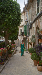 Fototapeta premium Young woman in a green dress strolling through picturesque valldemossa, mallorca, spain, enjoying a sunny day amidst beautiful stone buildings and vibrant potted plants