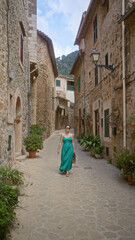 Woman walking down a picturesque narrow street in valldemossa, mallorca, spain, wearing a green dress and sunglasses, surrounded by stone buildings and lush plants.