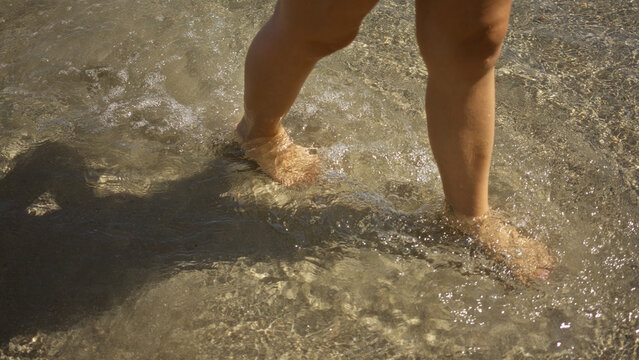 Woman strolling in shallow water at a sunny beach with her legs partially submerged, casting a shadow while enjoying the calm and clear ocean waves