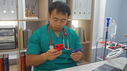 Young chinese male doctor in clinic holding smartphone and credit card, surrounded by medical...